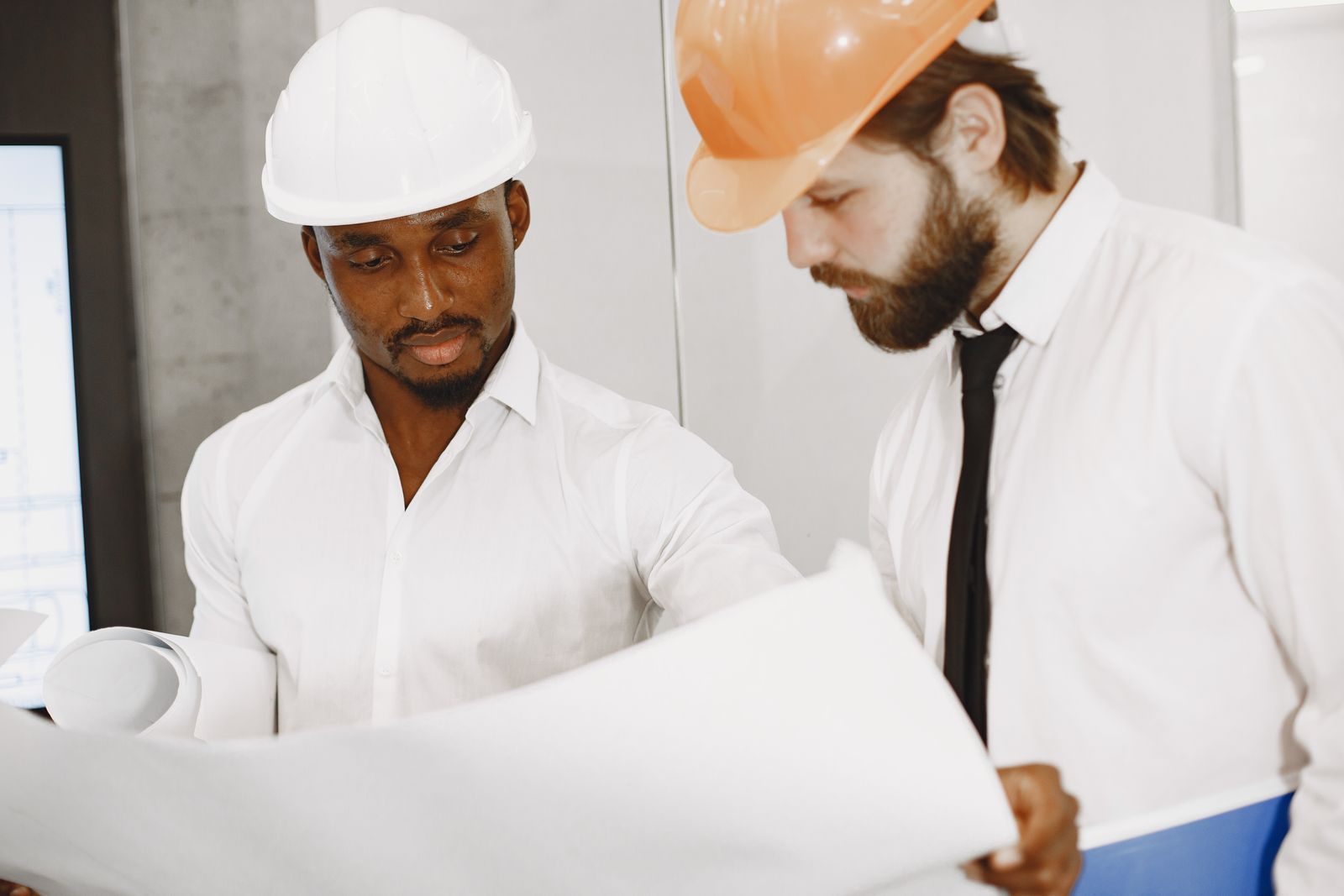 Two engineers in hard hats concentrate on reviewing detailed construction blueprints indoors